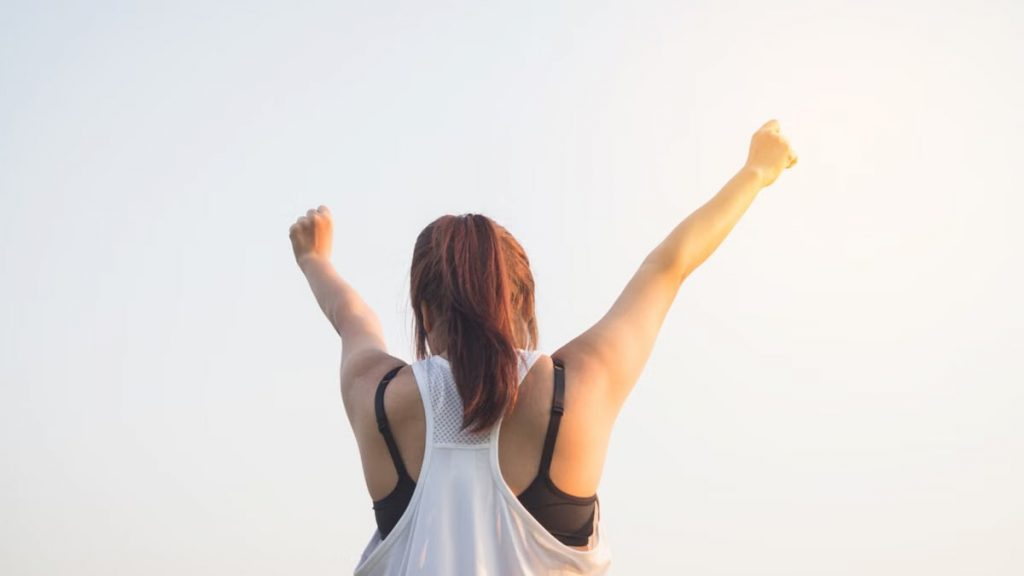 A woman celebrates success with arms raised in a bright outdoor setting.